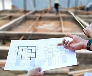 Person looking across a framed building, with a paper copy of building drawings in their hand.